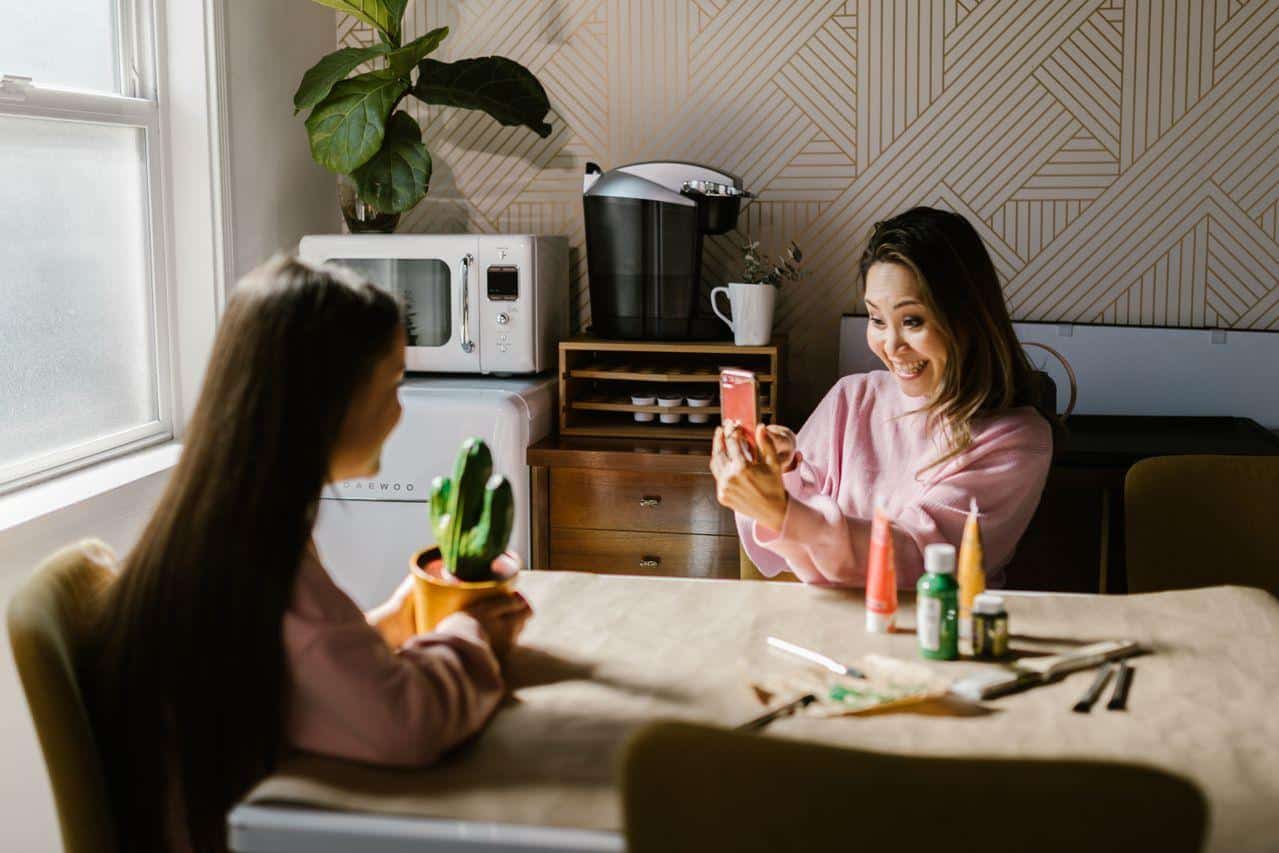 Mom smiling as she films her daughter holding a painted cactus pot at the kitchen table