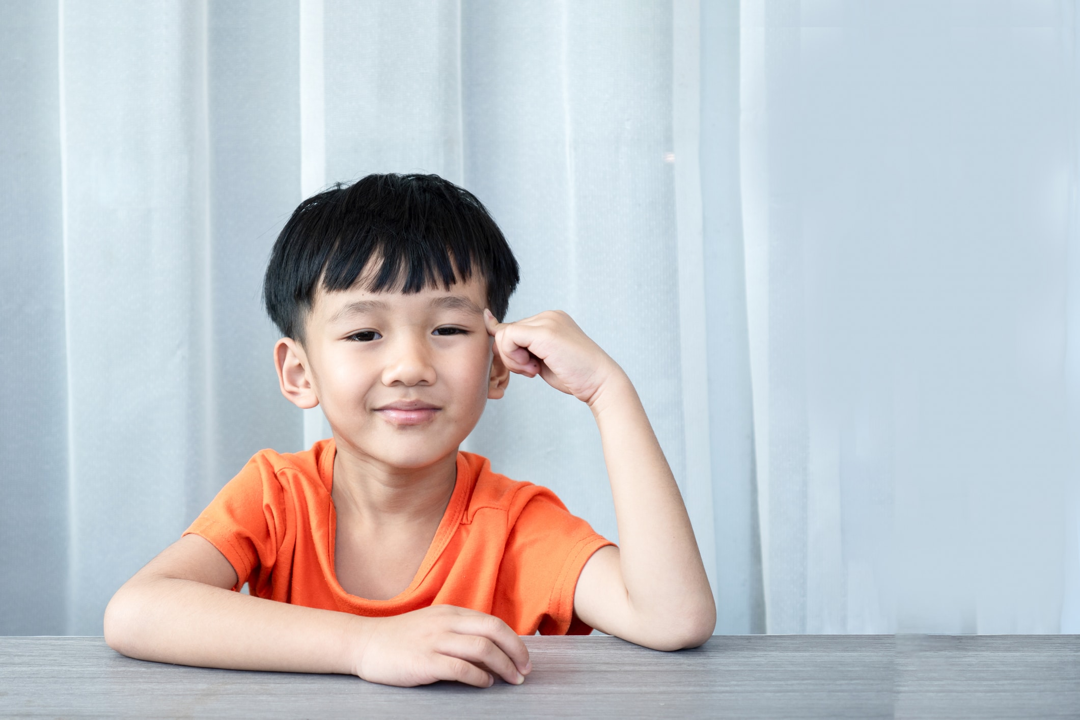 Young boy in an orange shirt resting his head on his hand while thinking at a table