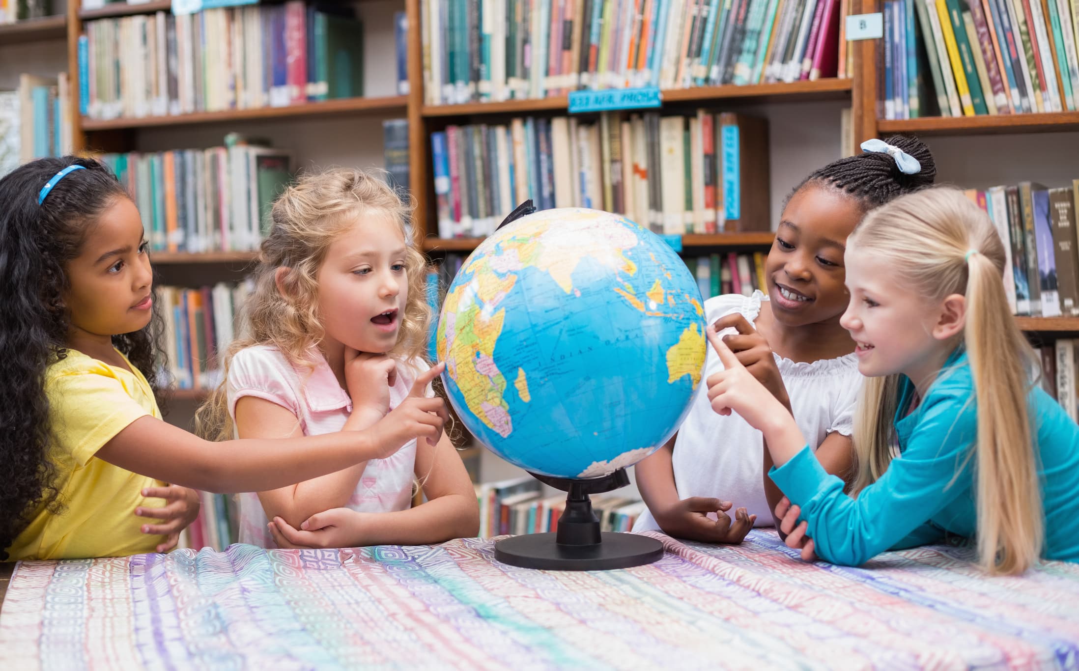 Four girls gathered around a globe in a school library, pointing at countries