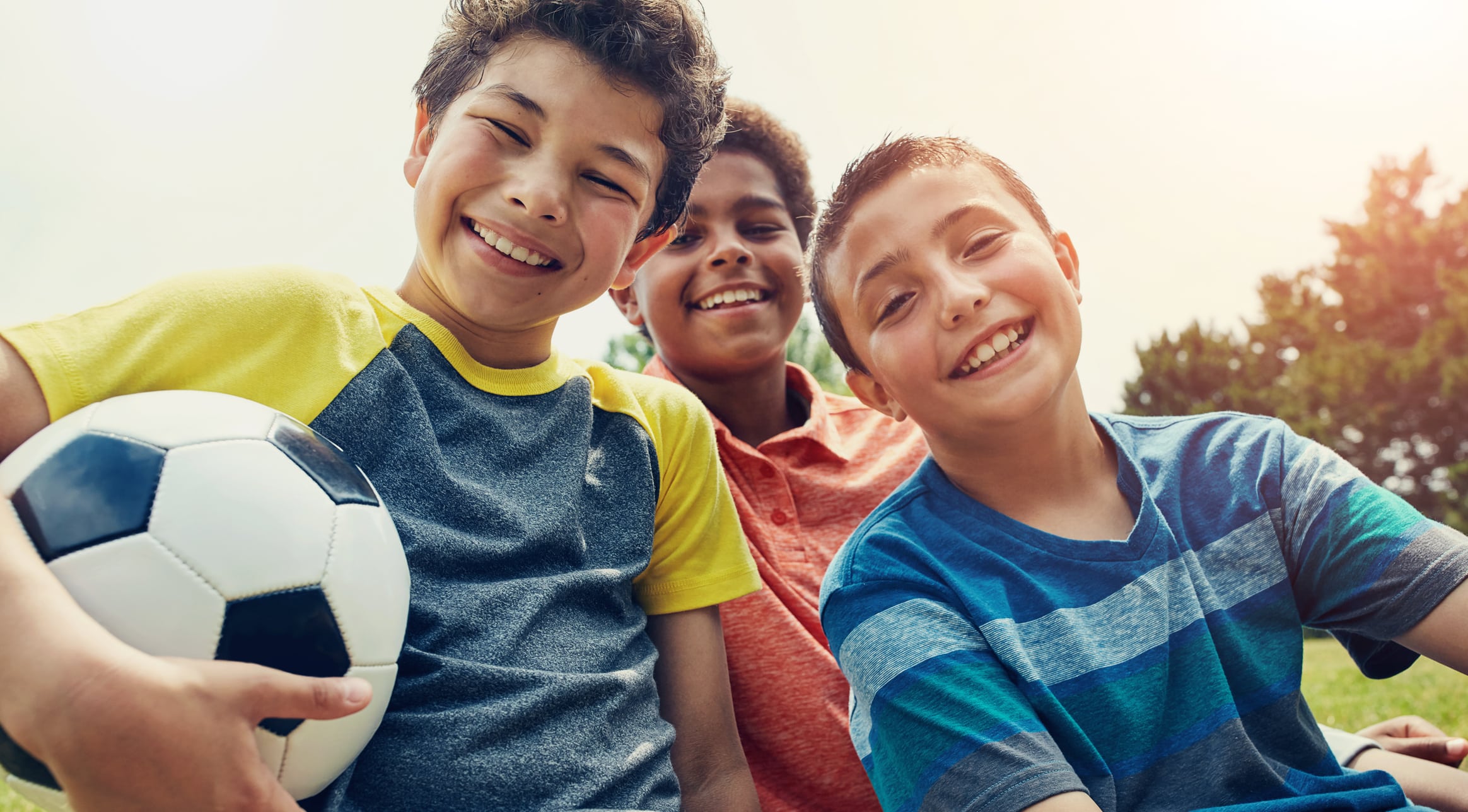Three smiling kids posing with a soccer ball in a park